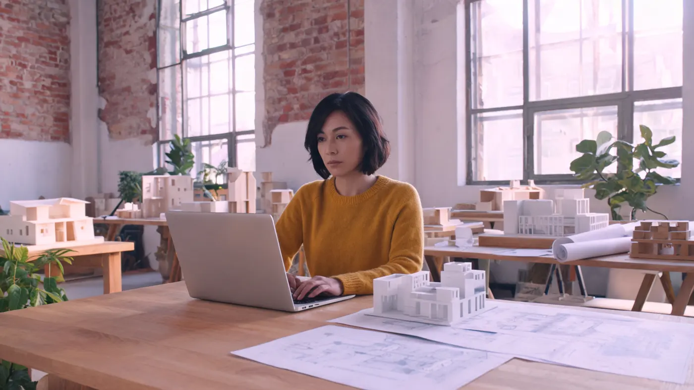 An architect writes on a laptop. A white building model is next to her.