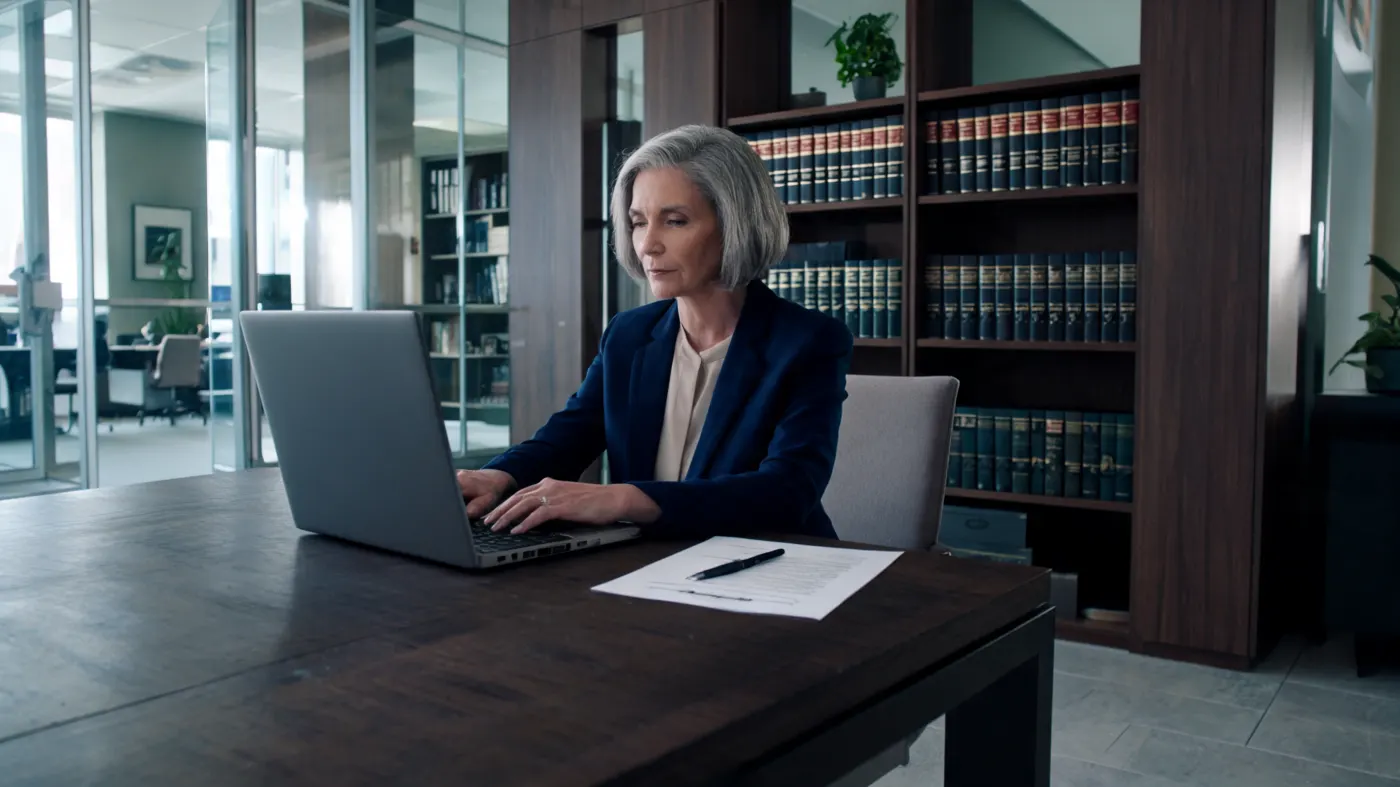 A lawyer writes a letter on a laptop. A printed contract is beside her.